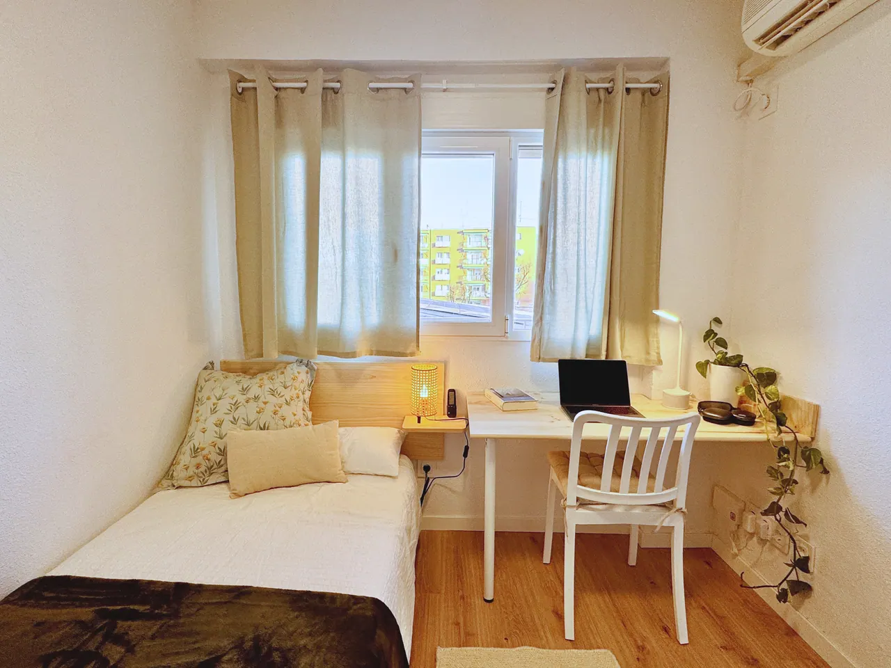 Habitación luminosa con cama individual, escritorio de madera, plantas y ventana con cortinas