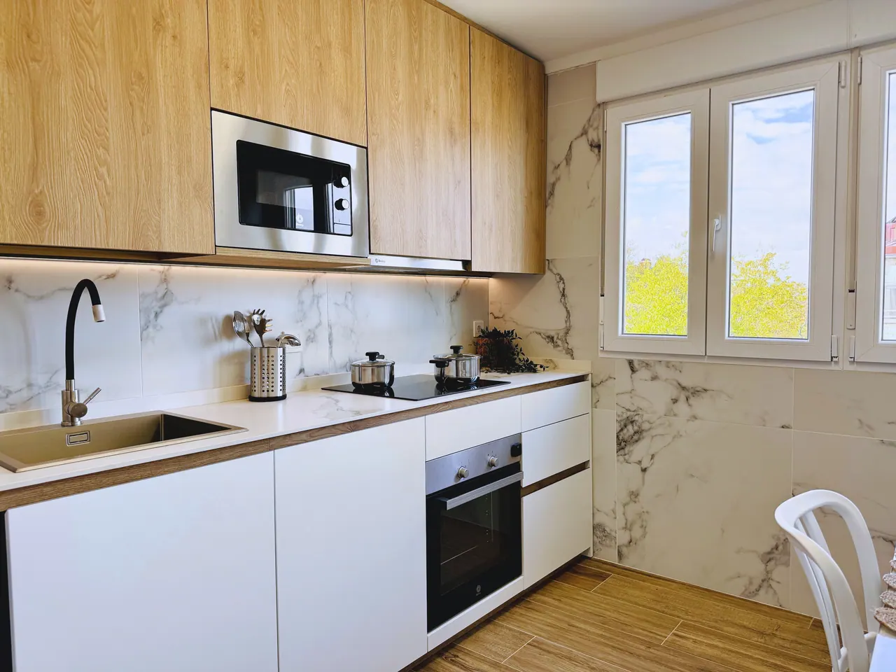 Cocina blanca con muebles altos de madera, pared de mármol, horno, microondas y ventana