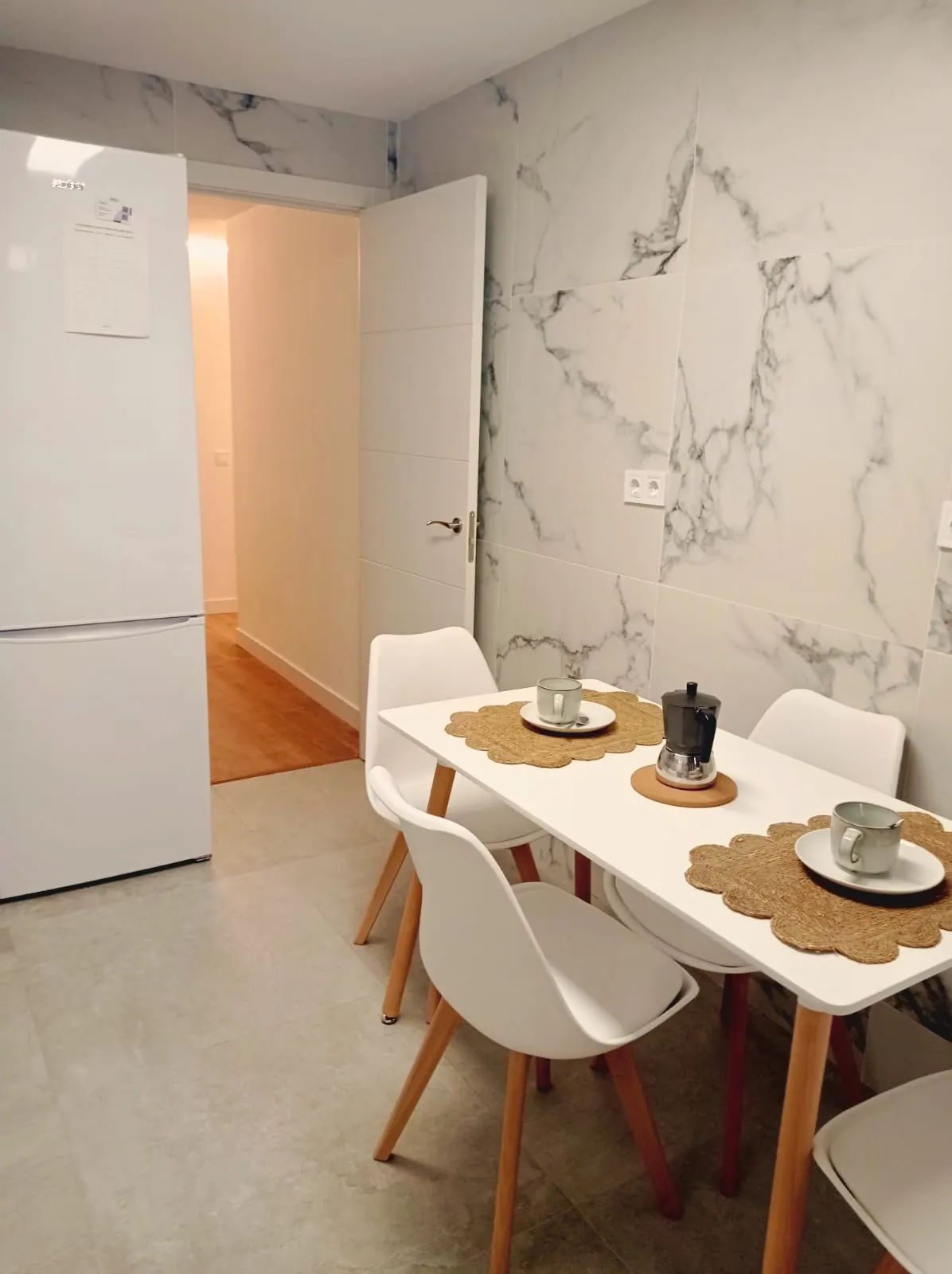 Mesa de comedor blanca con sillas y frigorífico junto a la pared de mármol