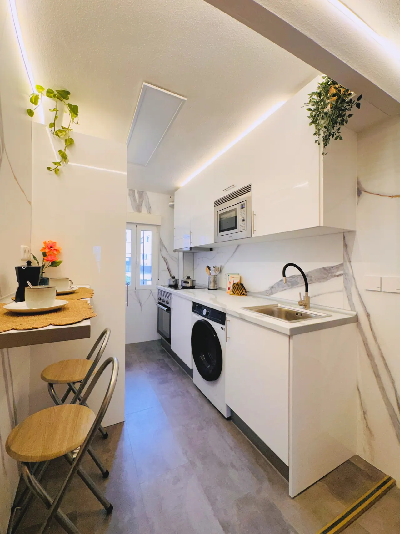 Cocina blanca alargada con lavadora, microondas, barra con taburetes y luz LED en el techo